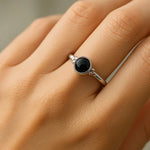 Eine Nahaufnahme einer Frauenhand zeigt einen eleganten Silberring mit einem runden, schwarzen Onyx-Cabochon. Der glatte, glänzende Stein steht im Kontrast zur polierten Ringschiene aus 925er Sterlingsilber. Weiches Licht betont die Details des Rings und die natürliche Textur der Haut, während der neutrale Hintergrund den Fokus auf das Schmuckstück lenkt.