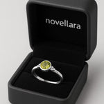 Ein silberner Ring mit rundem, hellgrünem Stein (Peridot-Optik) liegt in einer offenen, schwarzen Schmuckschatulle mit Samteinlage. Auf der Innenseite des Deckels steht das Logo „NOVELLARA“ in heller Schrift. Das Bild ist in Studioqualität vor hellem Hintergrund aufgenommen.