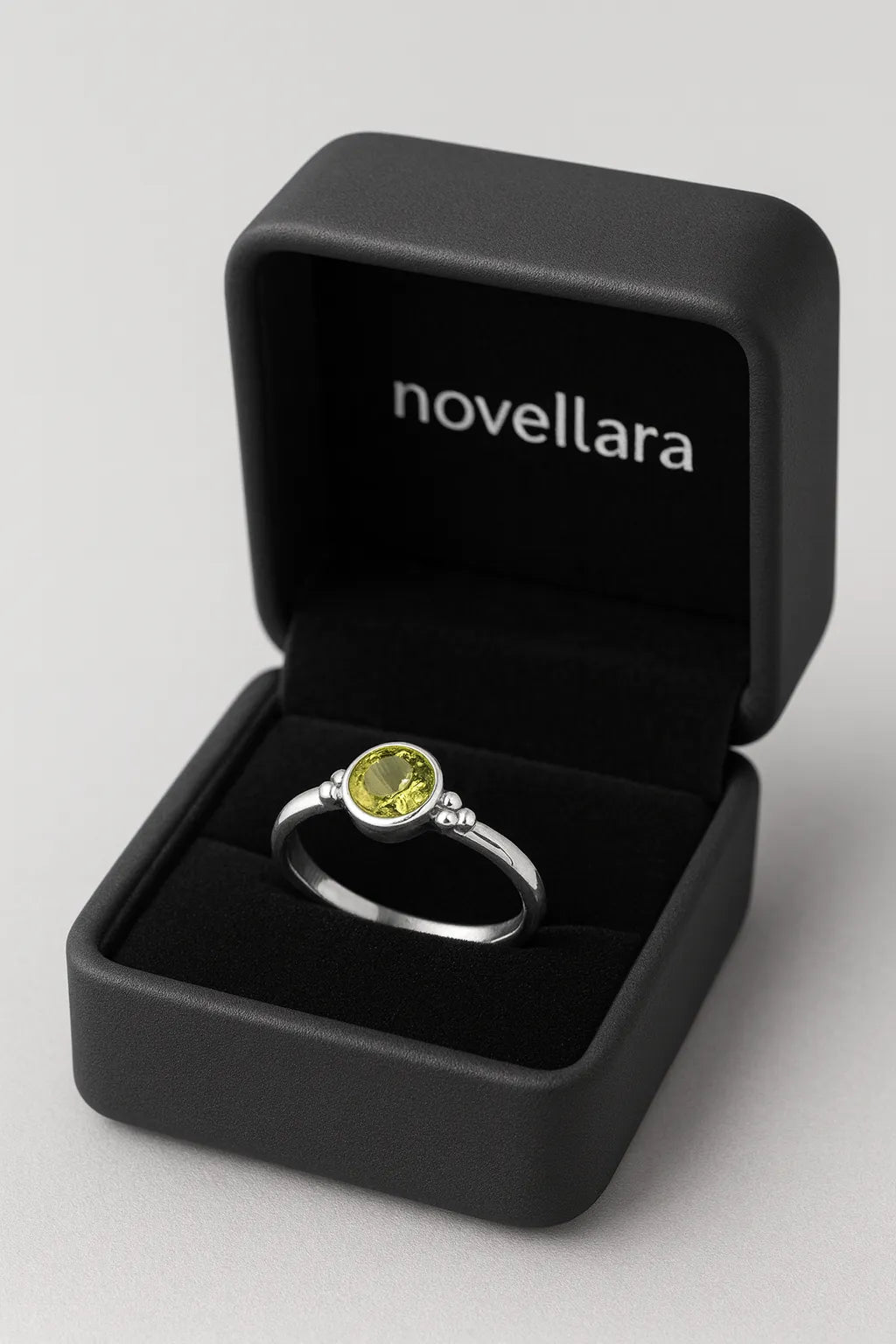 Ein silberner Ring mit rundem, hellgrünem Stein (Peridot-Optik) liegt in einer offenen, schwarzen Schmuckschatulle mit Samteinlage. Auf der Innenseite des Deckels steht das Logo „NOVELLARA“ in heller Schrift. Das Bild ist in Studioqualität vor hellem Hintergrund aufgenommen.