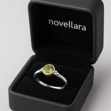 Ein silberner Ring mit rundem, hellgrünem Stein (Peridot-Optik) liegt in einer offenen, schwarzen Schmuckschatulle mit Samteinlage. Auf der Innenseite des Deckels steht das Logo „NOVELLARA“ in heller Schrift. Das Bild ist in Studioqualität vor hellem Hintergrund aufgenommen.