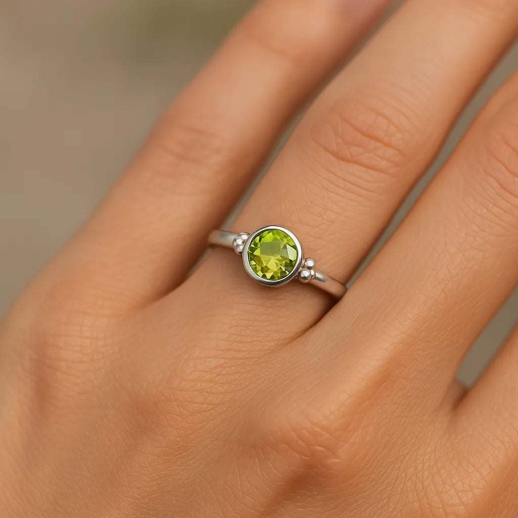 Eine Nahaufnahme einer Frauenhand zeigt einen silbernen Ring mit einem glatten, hellgrünen Peridot-Stein. Der Fokus liegt auf dem Ring, der im weichen, natürlichen Licht schimmert. Der Hintergrund ist neutral und unscharf, wodurch der edle Glanz des Silbers und die klare Farbe des Steins besonders betont werden.
