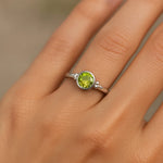Eine Nahaufnahme einer Frauenhand zeigt einen silbernen Ring mit einem glatten, hellgrünen Peridot-Stein. Der Fokus liegt auf dem Ring, der im weichen, natürlichen Licht schimmert. Der Hintergrund ist neutral und unscharf, wodurch der edle Glanz des Silbers und die klare Farbe des Steins besonders betont werden.