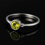 Feiner Silberring (gestempelt 925) mit rundem, facettiertem, leuchtend gelbgrünem Peridot in Zargenfassung und kleinen Silberkügelchen an den Seiten, liegend auf schwarzem, spiegelndem Hintergrund.