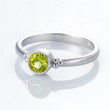 Schlichter Silberring mit einem runden, facettierten, hellgrün leuchtenden Stein (Peridot) in einer runden Fassung, seitlich mit kleinen silbernen Kügelchen verziert, auf weißem Hintergrund liegend und spiegelnd.