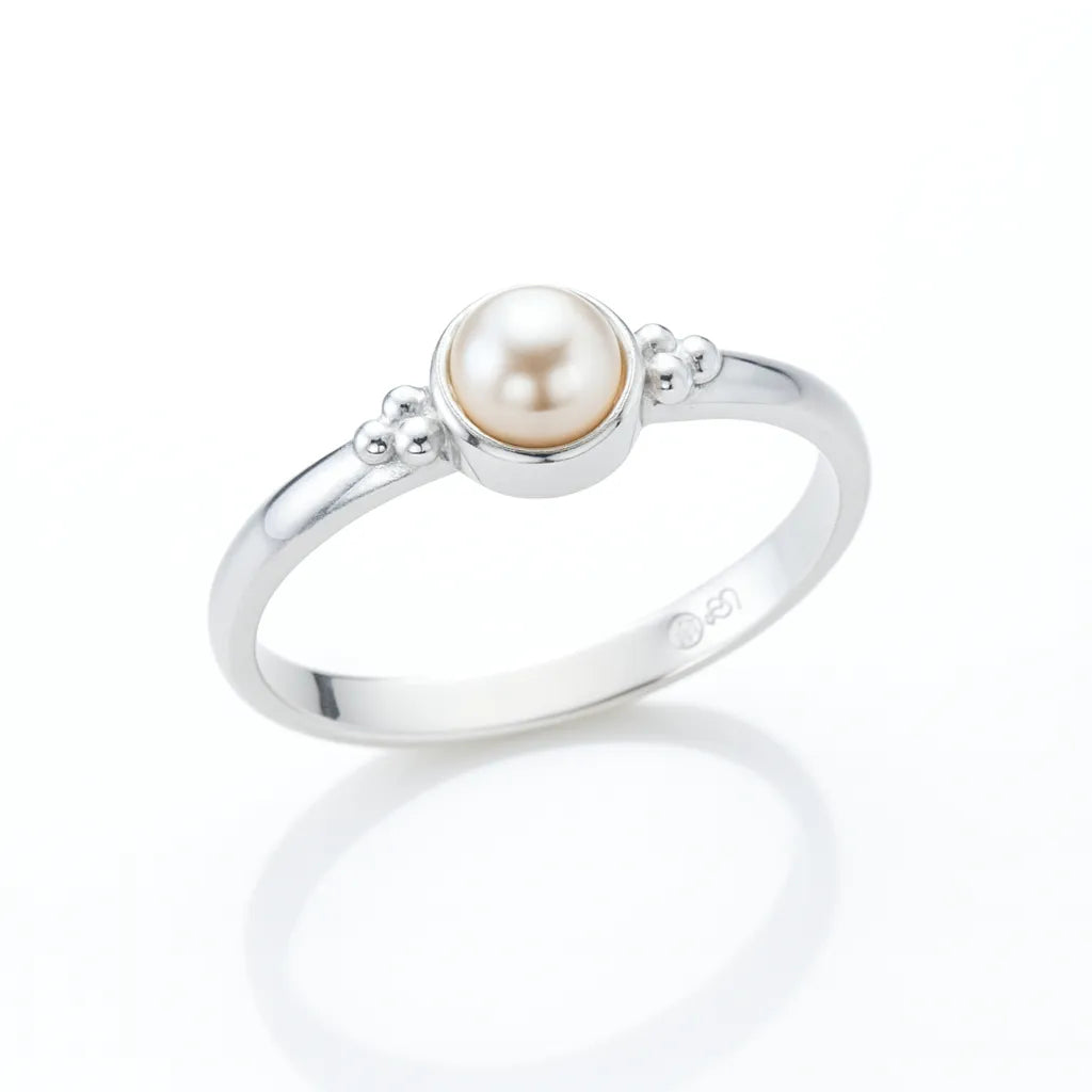 Eleganter Silberring mit runder Creme-Perle in Zargenfassung, seitlich fotografiert auf hellem spiegelnden Untergrund mit weicher Reflexion, filigrane Silberkügelchen als Detail am Ringkopf