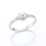 Eleganter Silberring mit runder Creme-Perle in Zargenfassung, seitlich fotografiert auf hellem spiegelnden Untergrund mit weicher Reflexion, filigrane Silberkügelchen als Detail am Ringkopf