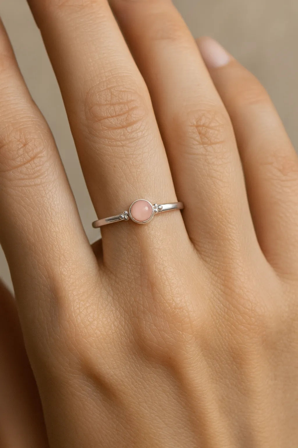 Nahaufnahme einer Frauenhand mit einem filigranen Silberring am Ringfinger. Der Ring ist mit einem kleinen, etwa 4 mm großen, runden Rosenquarz besetzt, der in einer glatten Zargenfassung sitzt. Der zart rosafarbene Stein wirkt leicht transparent und harmoniert mit dem silbernen Glanz des Rings. Die Haut ist warm getönt und das weiche Licht betont sowohl die feine Oberfläche des Rings als auch die natürliche Struktur der Haut.
