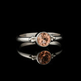 Eleganter Silberring mit rundem pfirsichfarbenem Zirkonia in Zargenfassung und kleinen Silberkügelchen, liegend auf schwarzem spiegelnden Untergrund mit starker Reflexion in hochwertiger Schmuck-Nahaufnahme.