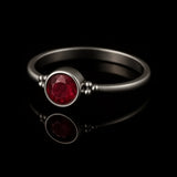 Eleganter Silberring mit rundem, facettierten Rubin in Zargenfassung und kleinen Silberkügelchen an den Seiten, liegend auf schwarzem, glänzendem Untergrund mit deutlicher Spiegelung des Rings.