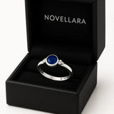Ein silberner Ring mit einem runden, blauen 4-mm-Stein liegt in einer geöffneten schwarzen Schmuckschatulle mit samtigem Inlay. Auf der Innenseite des Deckels steht das Logo „NOVELLARA“ in heller Schrift. Das Bild ist in Studioqualität vor hellem Hintergrund aufgenommen.