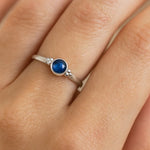 Nahaufnahme einer Frauenhand mit einem eleganten Silberring am Ringfinger. Der Ring trägt einen etwa 4 mm großen, facettierten blauen Saphir in einer runden Zargenfassung, flankiert von kleinen Silberkügelchen auf beiden Seiten. Das Licht betont die Tiefe und Brillanz des Saphirs sowie den dezenten Glanz des polierten Silbers, während die natürliche Struktur der Haut weich und realistisch zur Geltung kommt.