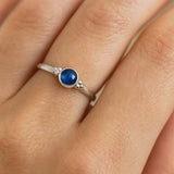 Nahaufnahme einer Frauenhand mit einem eleganten Silberring am Ringfinger. Der Ring trägt einen etwa 4 mm großen, facettierten blauen Saphir in einer runden Zargenfassung, flankiert von kleinen Silberkügelchen auf beiden Seiten. Das Licht betont die Tiefe und Brillanz des Saphirs sowie den dezenten Glanz des polierten Silbers, während die natürliche Struktur der Haut weich und realistisch zur Geltung kommt.
