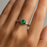 Ein eleganter Silberring mit einem runden, grünen Smaragd-Cabochon, der an der Hand einer Frau getragen wird. Der Fokus liegt auf dem Ring, der durch seine glatte, polierte Fassung und den leuchtend grünen Stein in 4 mm Größe hervorsticht. Die Szene ist neutral ausgeleuchtet und betont die feine Handarbeit des Schmuckstücks.