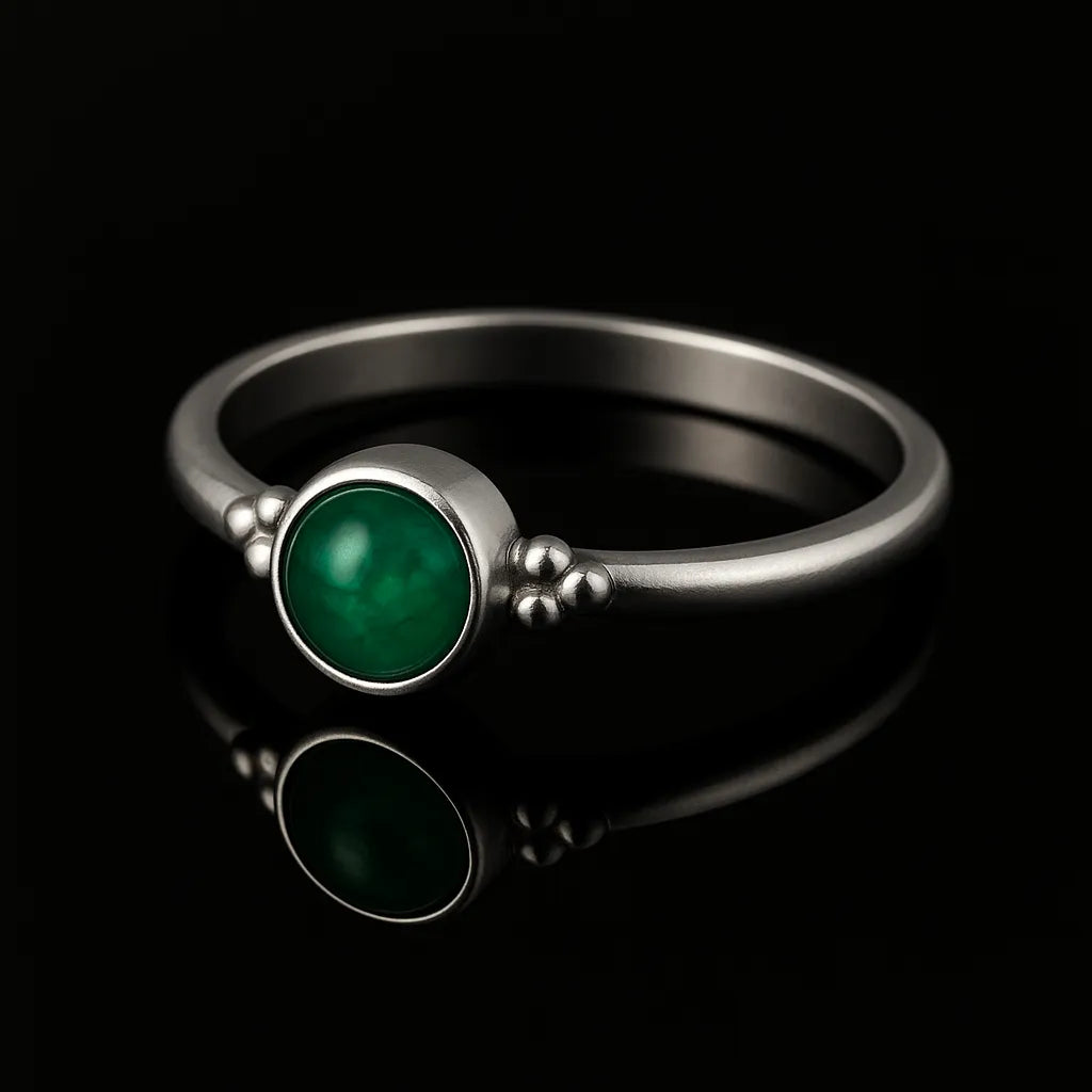 Schlichter Silberring mit rundem, glatten, smaragdgrün leuchtenden Stein in Zargenfassung und kleinen Silberkügelchen an den Seiten, liegend auf schwarzem, spiegelndem Untergrund mit deutlicher Reflexion des Rings.