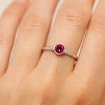 Nahaufnahme einer Frauenhand mit einem filigranen Silberring am Ringfinger. Der Ring besitzt einen kleinen, etwa 4 mm großen, facettierten roten Turmalin in einer runden Zargenfassung. Neben dem Stein befinden sich zwei kleine Silberkügelchen auf jeder Seite. Das weiche Licht hebt die glänzende Oberfläche des Silbers, die klare Facettierung des Edelsteins und die natürliche Struktur der Haut hervor.