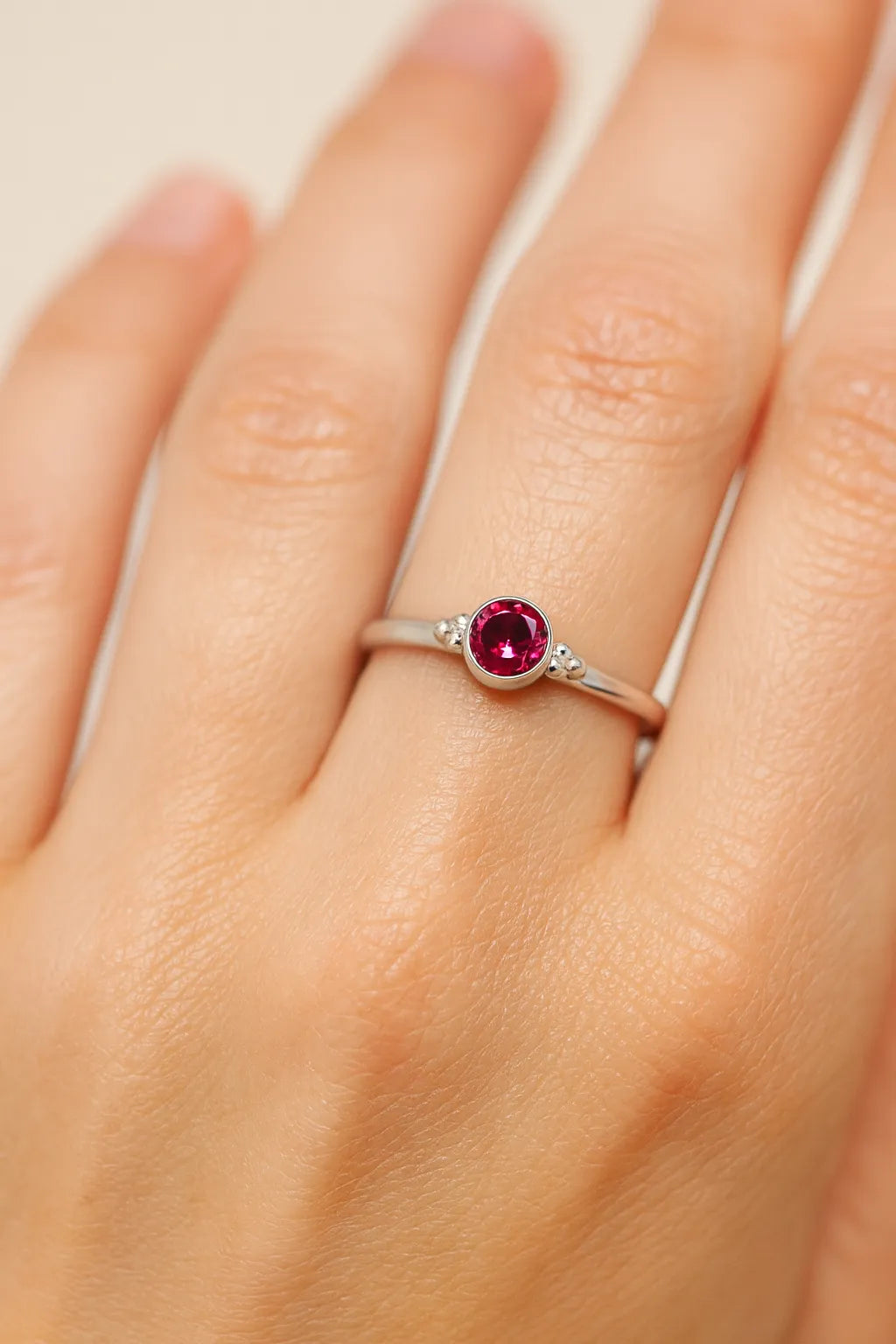 Nahaufnahme einer Frauenhand mit einem filigranen Silberring am Ringfinger. Der Ring besitzt einen kleinen, etwa 4 mm großen, facettierten roten Turmalin in einer runden Zargenfassung. Neben dem Stein befinden sich zwei kleine Silberkügelchen auf jeder Seite. Das weiche Licht hebt die glänzende Oberfläche des Silbers, die klare Facettierung des Edelsteins und die natürliche Struktur der Haut hervor.