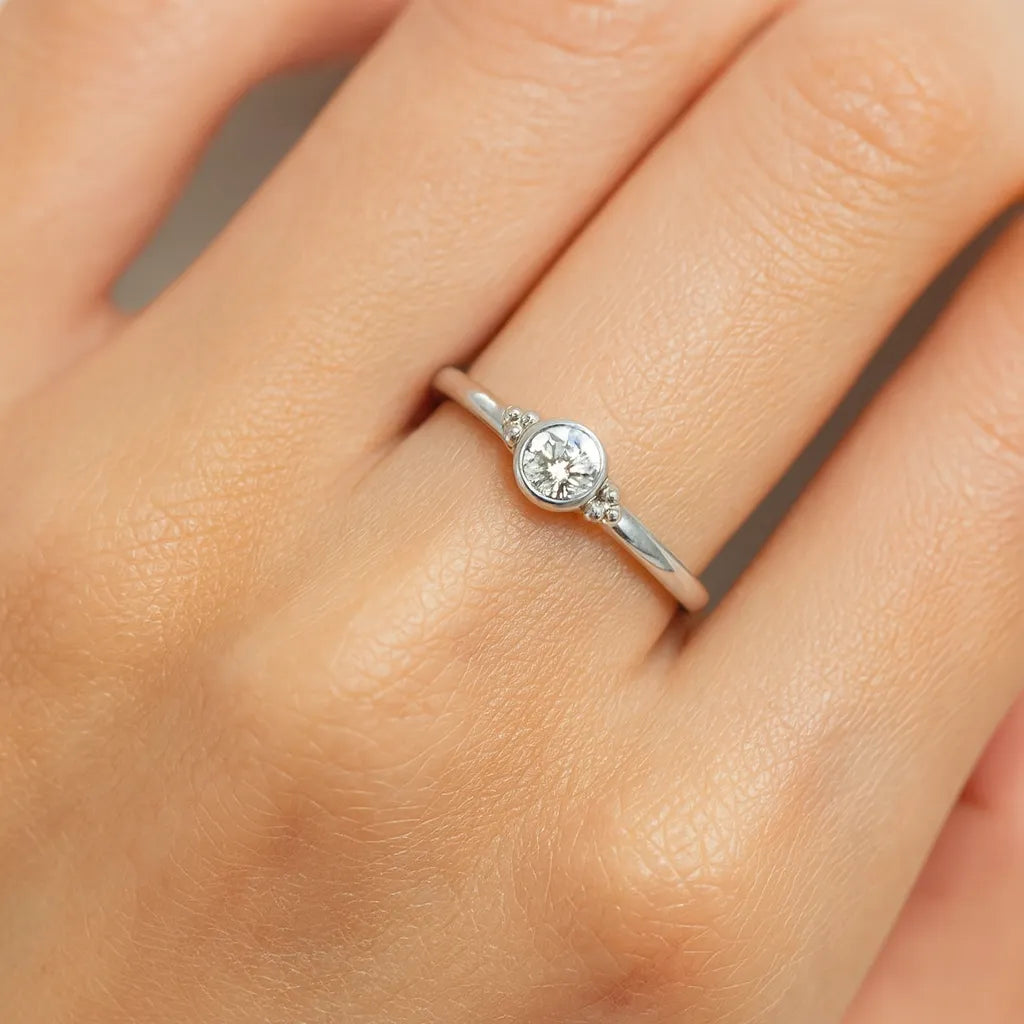 Eine Nahaufnahme einer Frauenhand zeigt einen eleganten Silberring am Ringfinger. Der Ring besitzt ein schlichtes, rundes Design mit einem klar facettierten Zirkonia-Stein in der Mitte, der in einer glatten Fassung eingefasst ist. Der Fokus des Bildes liegt auf dem Ring, der auf der Haut leicht glänzt und das Licht fein reflektiert.