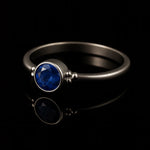 Eleganter Silberring mit rundem, facettierten, intensiv königsblauen Stein in Zargenfassung und dekorativen Silberkügelchen an den Seiten, liegend auf schwarzem, stark reflektierendem Untergrund mit klarer Spiegelung des Rings.