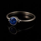 Eleganter Silberring mit rundem, facettierten, intensiv königsblauen Stein in Zargenfassung und dekorativen Silberkügelchen an den Seiten, liegend auf schwarzem, stark reflektierendem Untergrund mit klarer Spiegelung des Rings.