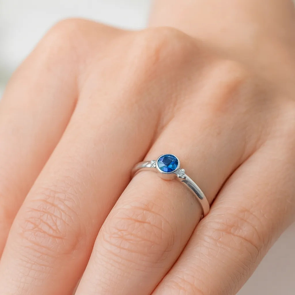 Nahaufnahme einer Frauenhand mit einem eleganten Silberring am Ringfinger. Der Ring besitzt einen runden, facettierten blauen Kyanit-Edelstein in einer schlichten Zargenfassung, flankiert von kleinen Silberkügelchen. Die Haut ist natürlich und leicht warm getönt, und das weiche Licht hebt den Glanz des Rings sowie die feinen Facetten des Steins hervor.