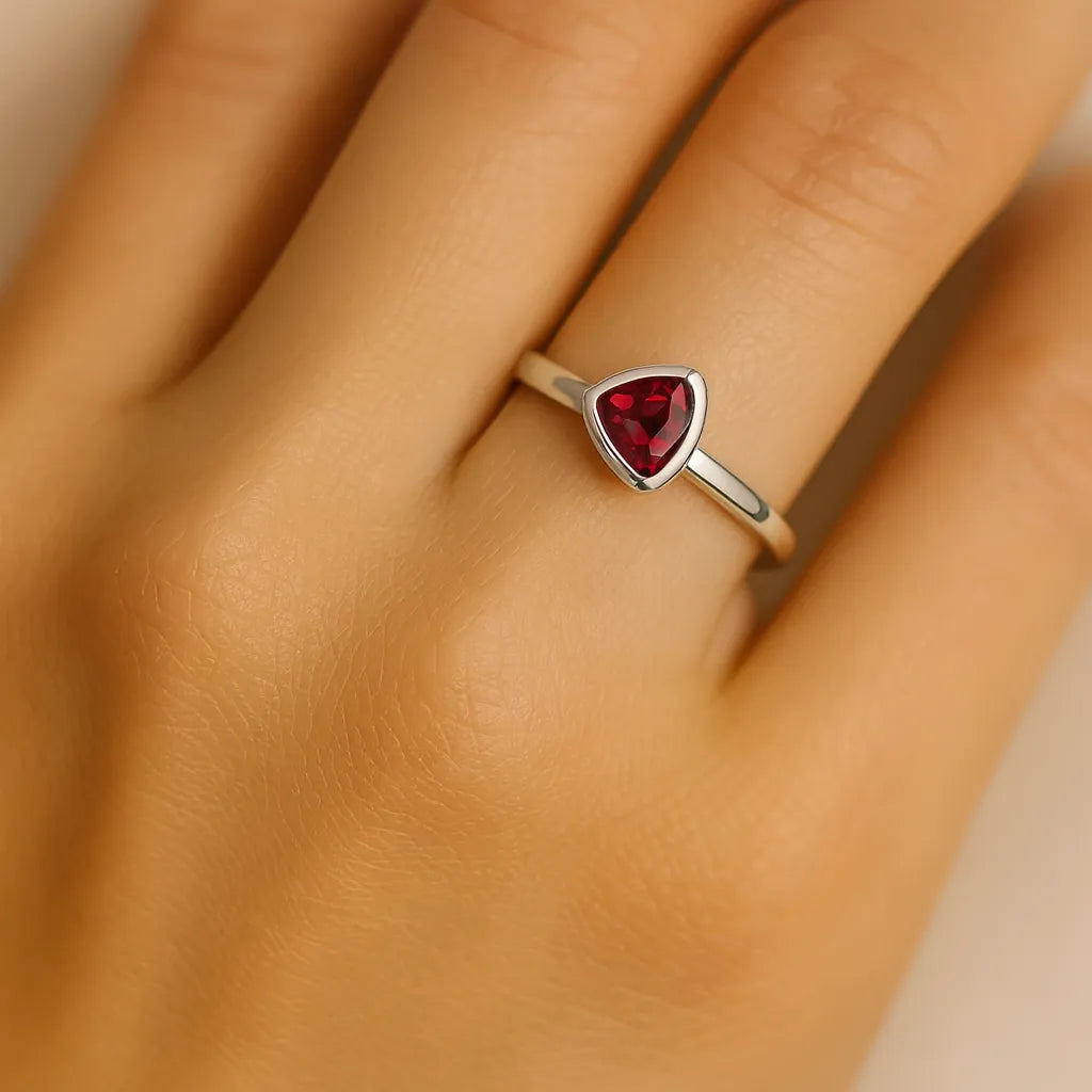 Nahaufnahme einer Frauenhand, die einen eleganten Silberring mit einem dreieckig geschliffenen roten Granat auf dem Ringfinger trägt. Der Fokus liegt auf dem Ring, dessen polierte Fassung den glänzenden Edelstein betont. Die Haut wirkt weich und natürlich beleuchtet, wodurch der Ring besonders hervorgehoben wird.