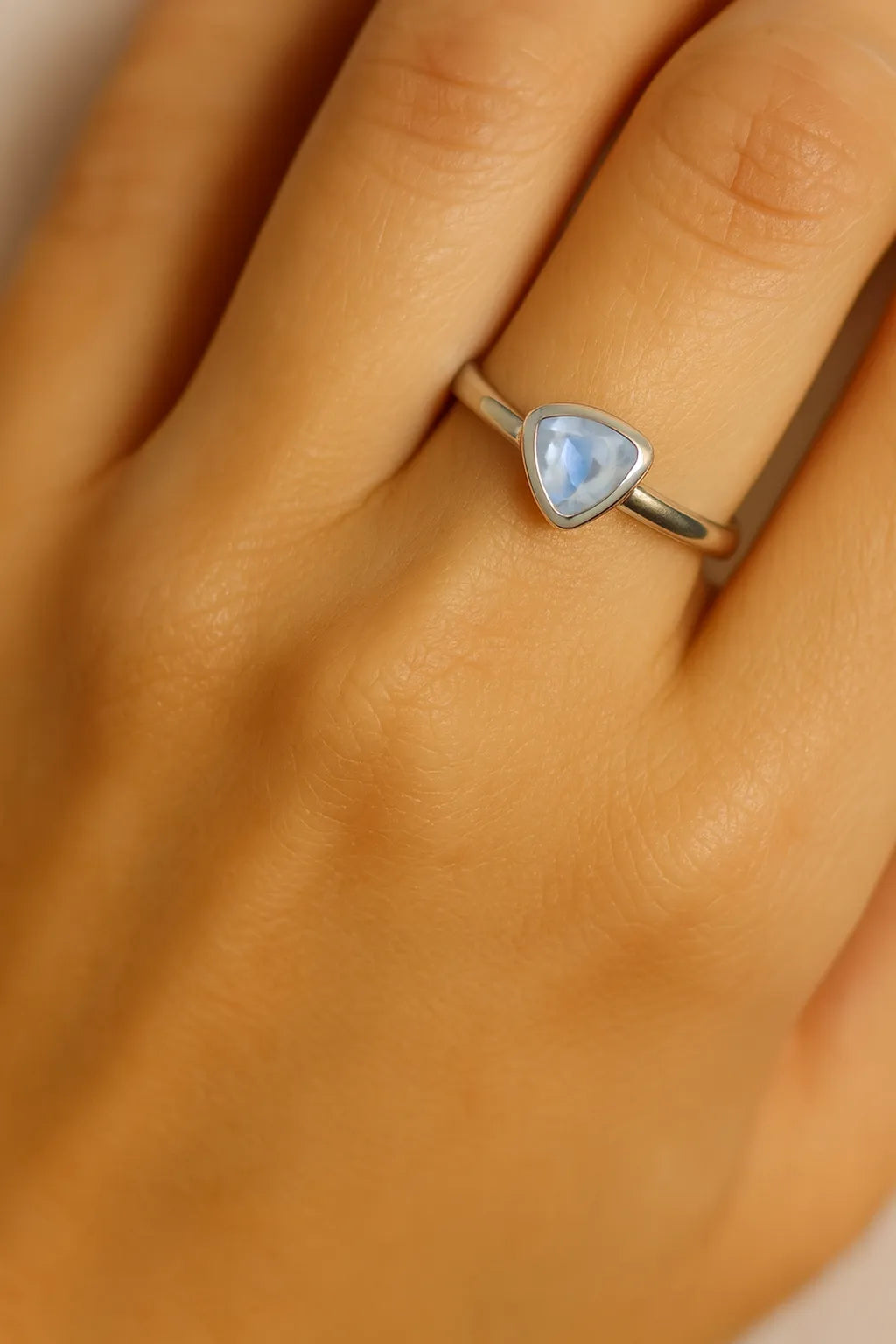 Eine Nahaufnahme einer Frauenhand zeigt einen eleganten Silberring am Ringfinger. Der Ring besitzt eine schlichte, glatte Ringschiene und ist mit einem dreieckig facettierten, hellblauen Blautopas von etwa 5 mm Länge besetzt. Der Fokus liegt klar auf dem Ring und seinem funkelnden Edelstein, während die Haut im Hintergrund weich und natürlich wirkt.