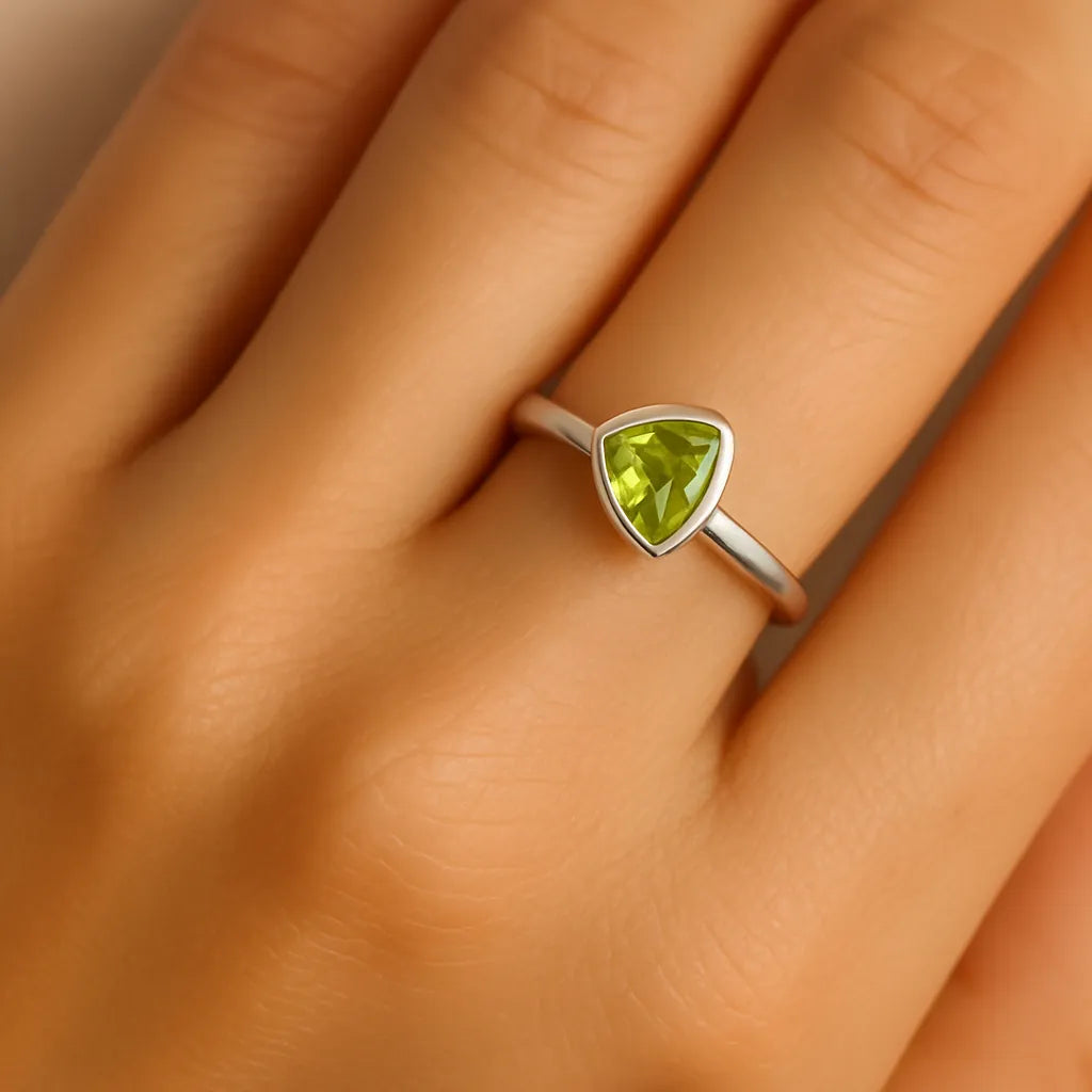 Ein realistisches Foto zeigt die linke Hand einer Frau, die einen eleganten Silberring mit einem dreieckig geschliffenen, hellgrünen Peridot-Stein am Ringfinger trägt. Der Ring hat eine schlichte, glatte Ringschiene und eine dezente Fassung, die den 5 mm großen Stein sicher hält. Der Fokus liegt klar auf dem Ring, während der Hautton natürlich und weich beleuchtet ist.
