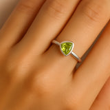 Ein realistisches Foto zeigt die linke Hand einer Frau, die einen eleganten Silberring mit einem dreieckig geschliffenen, hellgrünen Peridot-Stein am Ringfinger trägt. Der Ring hat eine schlichte, glatte Ringschiene und eine dezente Fassung, die den 5 mm großen Stein sicher hält. Der Fokus liegt klar auf dem Ring, während der Hautton natürlich und weich beleuchtet ist.