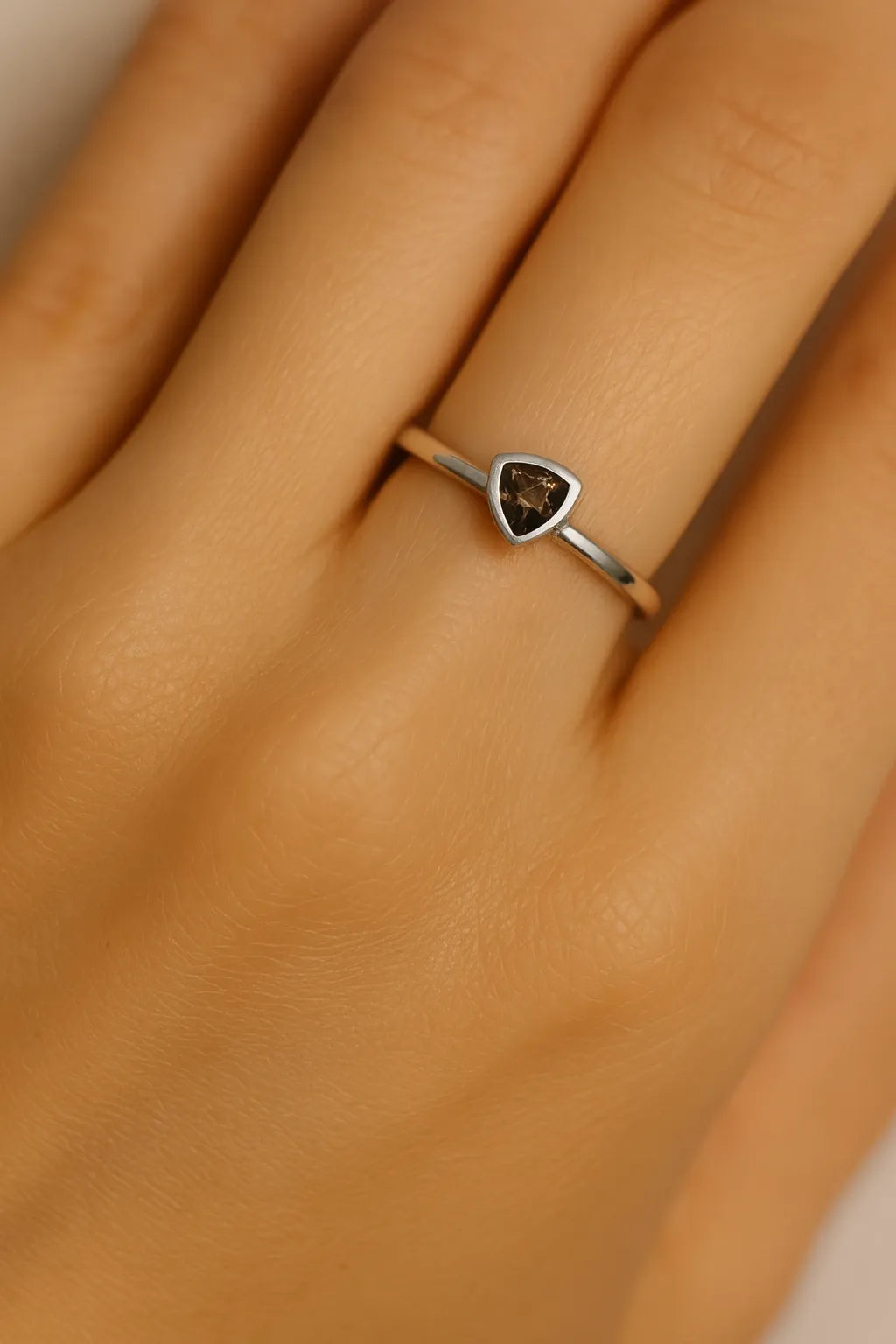 Nahaufnahme einer Frauenhand mit einem eleganten Silberring am Ringfinger. Der Ring trägt einen dreieckig facettierten Rauchquarz in warmer, brauner Farbe, der in einer glatten Fassung eingefasst ist. Der Fokus liegt klar auf dem Ring und dem glänzenden Edelstein, während der Hintergrund sanft verschwimmt.