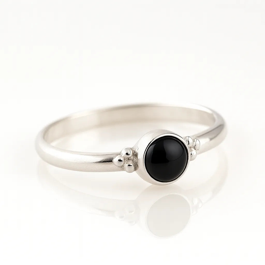 Feiner Silberring mit rundem schwarzem Onyx-Cabochon in Zargenfassung, liegend auf hellem spiegelnden Untergrund mit sanfter Reflexion, moderne seitliche Produktaufnahme für Damenschmuck.
