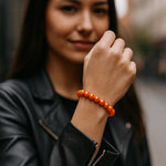 Eine junge Frau trägt ein orangefarbenes Perlenarmband am Handgelenk. Der Fokus liegt auf dem Armband, während sie in einer urbanen Umgebung steht und eine schwarze Lederjacke trägt.