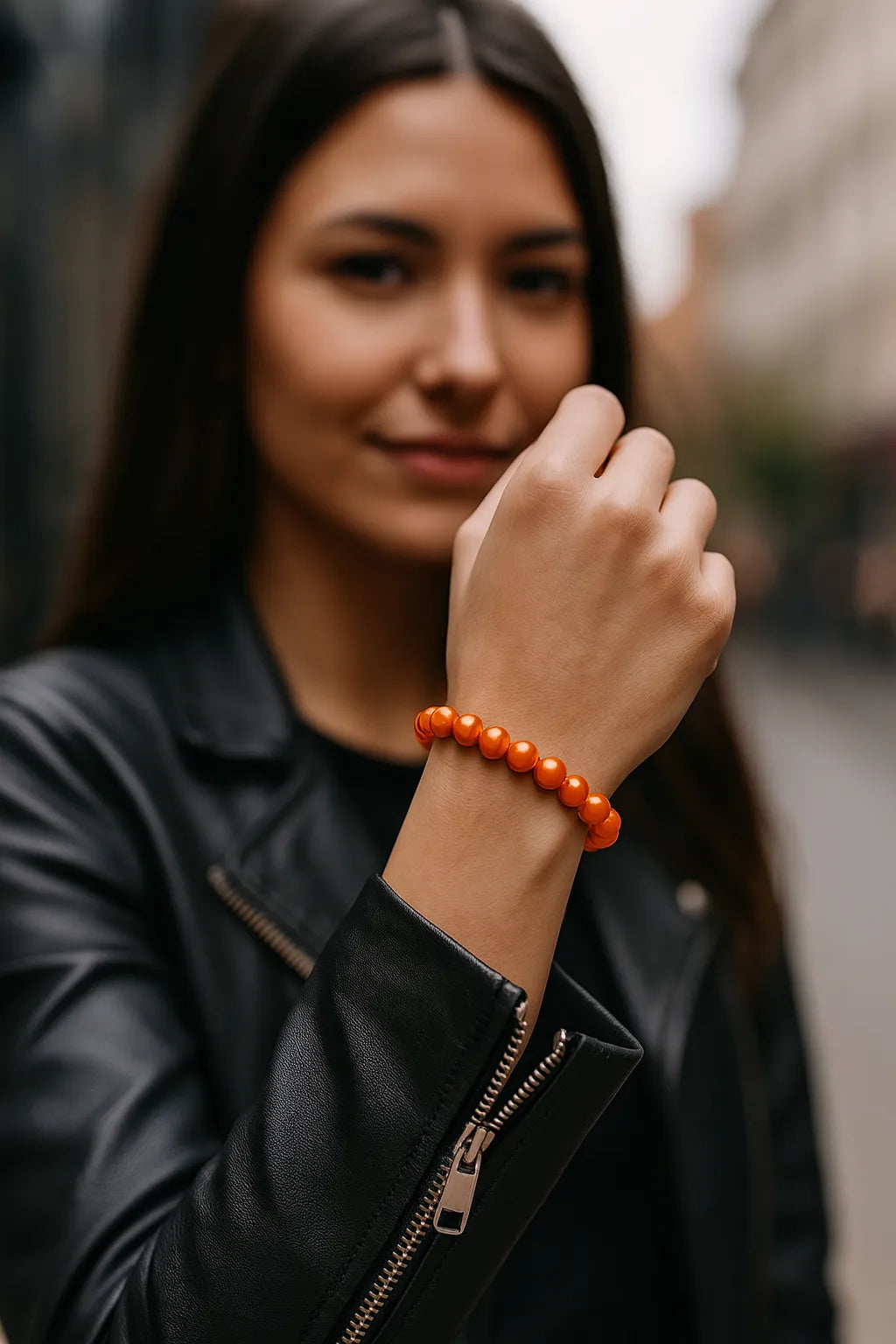 Eine junge Frau trägt ein orangefarbenes Perlenarmband am Handgelenk. Der Fokus liegt auf dem Armband, während sie in einer urbanen Umgebung steht und eine schwarze Lederjacke trägt.