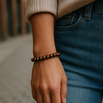 Eine Person trägt ein bronzefarbenes Perlenarmband am Handgelenk. Der Fokus liegt auf dem Armband, im Hintergrund ist eine unscharfe urbane Straßenszene erkennbar.