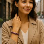 Eine Nahaufnahme einer Frau mit braunen Haaren, die eine beige Jacke und ein weißes Oberteil trägt und eine lila Perlenarmkette am linken Handgelenk hat. Sie lächelt leicht und ihr rechter Arm ist verschränkt, während ihre Hand ihren Hals berührt. Der Hintergrund ist leicht unscharf und zeigt eine städtische Umgebung mit Gebäuden und Bäumen.