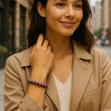 Eine Nahaufnahme einer Frau mit braunen Haaren, die eine beige Jacke und ein weißes Oberteil trägt und eine lila Perlenarmkette am linken Handgelenk hat. Sie lächelt leicht und ihr rechter Arm ist verschränkt, während ihre Hand ihren Hals berührt. Der Hintergrund ist leicht unscharf und zeigt eine städtische Umgebung mit Gebäuden und Bäumen.