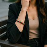 Eine modern gekleidete Frau sitzt auf einer dunklen Parkbank in urbaner Umgebung. Sie trägt ein schwarzes Sakko über einem beigefarbenen Top, und an ihrem Handgelenk befindet sich ein glänzendes Armband mit abwechselnd pinken und grünen Perlen. Der Fokus liegt auf dem Armband, während Hintergrund und restliche Szene sanft verschwommen sind.