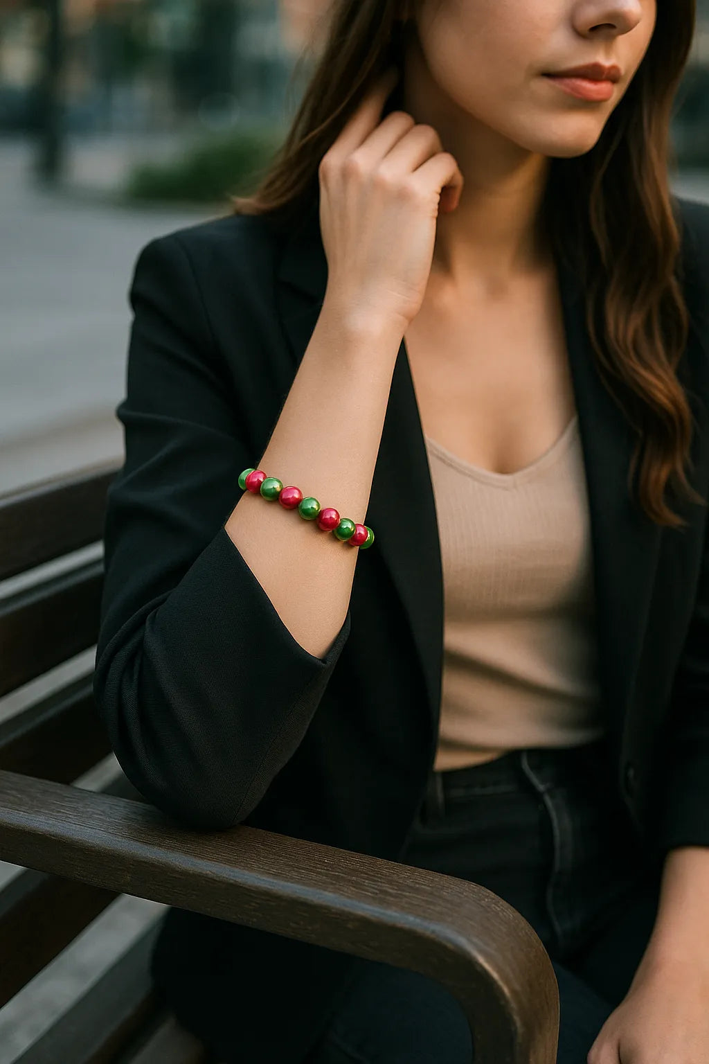 Eine modern gekleidete Frau sitzt auf einer dunklen Parkbank in urbaner Umgebung. Sie trägt ein schwarzes Sakko über einem beigefarbenen Top, und an ihrem Handgelenk befindet sich ein glänzendes Armband mit abwechselnd pinken und grünen Perlen. Der Fokus liegt auf dem Armband, während Hintergrund und restliche Szene sanft verschwommen sind.