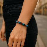 Eine Person trägt ein Armband aus abwechselnd türkisblauen und dunkelvioletten Perlen am Handgelenk. Sie steht auf einer städtischen Straße, trägt ein schwarzes Oberteil und schwarze Jeans. Der Fokus liegt auf dem Armband und dem Unterarm.