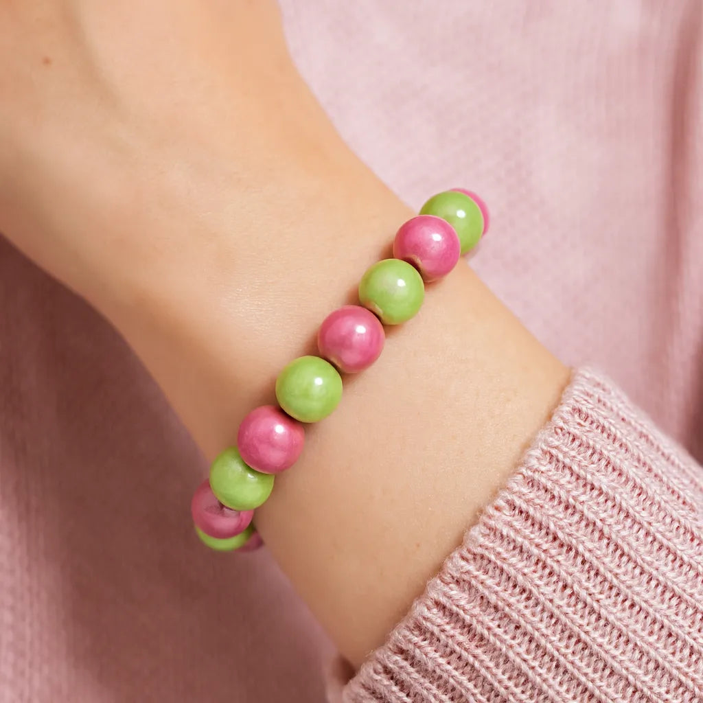 Nahaufnahme eines Handgelenks, das ein Armband aus abwechselnd pinken und grünen Perlen trägt. Die Person trägt einen hellrosa Strickpullover.