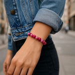 Eine Person trägt ein pink-rosa Perlenarmband am Handgelenk, während sie auf einer städtischen Straße steht. Sie trägt eine hochgekrempelte Jeansjacke und dunkle Hose. Der Hintergrund ist leicht unscharf, das Armband steht im Fokus.