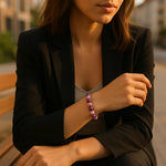 Eine modern gekleidete Frau sitzt auf einer Holzbank in einer städtischen Umgebung. Sie trägt ein schwarzes Sakko über einem beigen Top, und der Fokus liegt auf ihrem Handgelenk, an dem ein Armband aus abwechselnd rosa und lila glänzenden Perlen schimmert. Warmes Abendlicht betont die sanften Farben und schafft eine ruhige, elegante Atmosphäre.