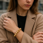 Eine Frau trägt ein goldenes Perlenarmband am Handgelenk. Sie steht in urbaner Umgebung, trägt einen beigefarbenen Mantel über einem schwarzen Oberteil. Der Fokus liegt auf dem Armband.