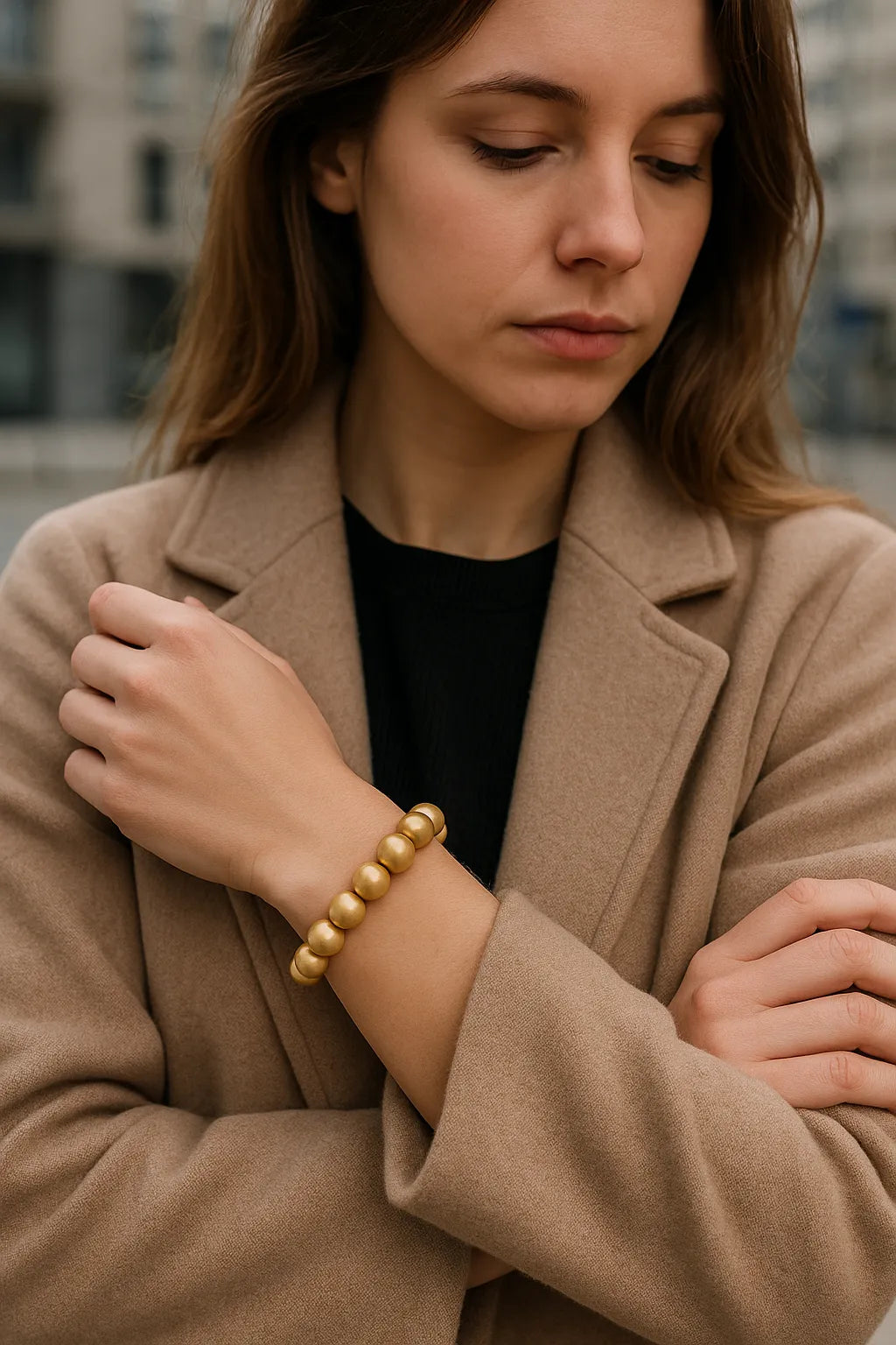 Eine Frau trägt ein goldenes Perlenarmband am Handgelenk. Sie steht in urbaner Umgebung, trägt einen beigefarbenen Mantel über einem schwarzen Oberteil. Der Fokus liegt auf dem Armband.