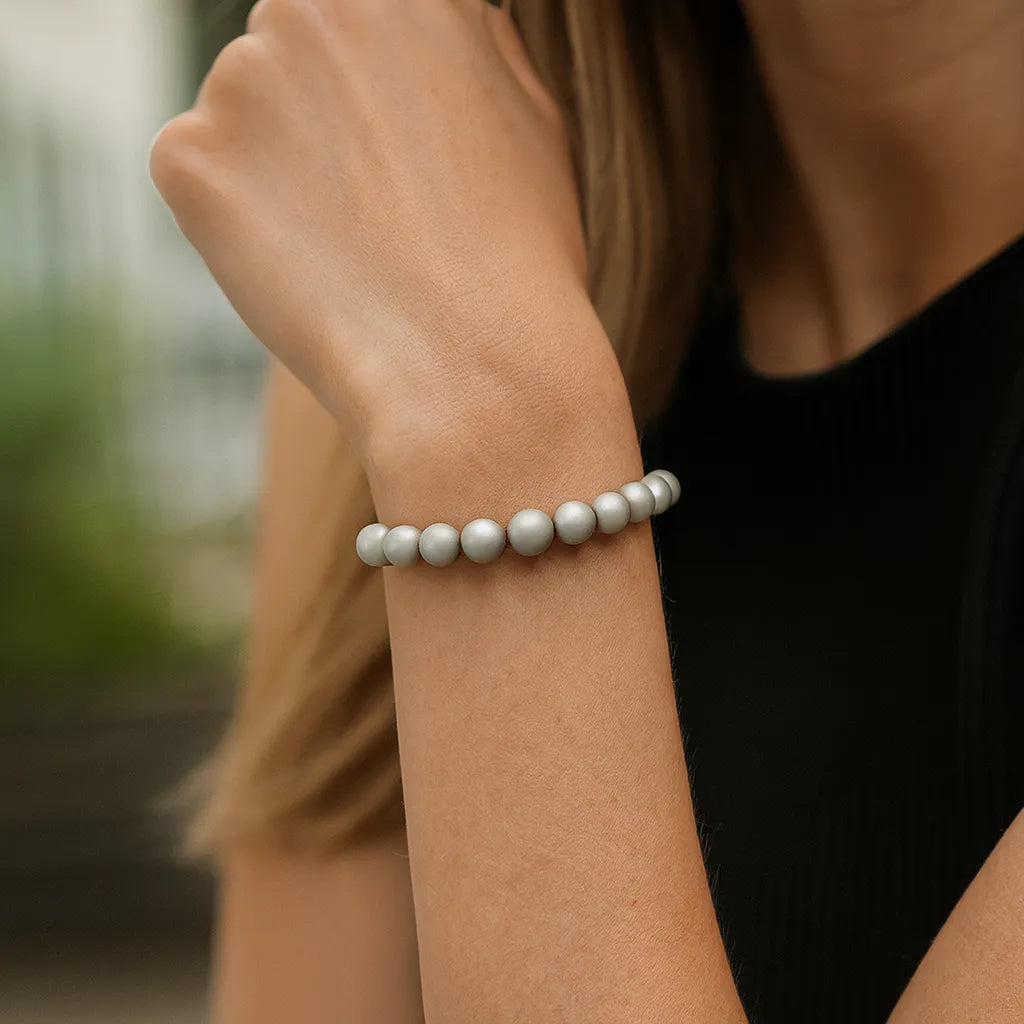 Eine Frau trägt ein silbernes Perlenarmband am Handgelenk. Der Fokus liegt auf dem Armband, während ihr Arm und ein schwarzes Oberteil im Vordergrund zu sehen sind. Der Hintergrund ist unscharf.