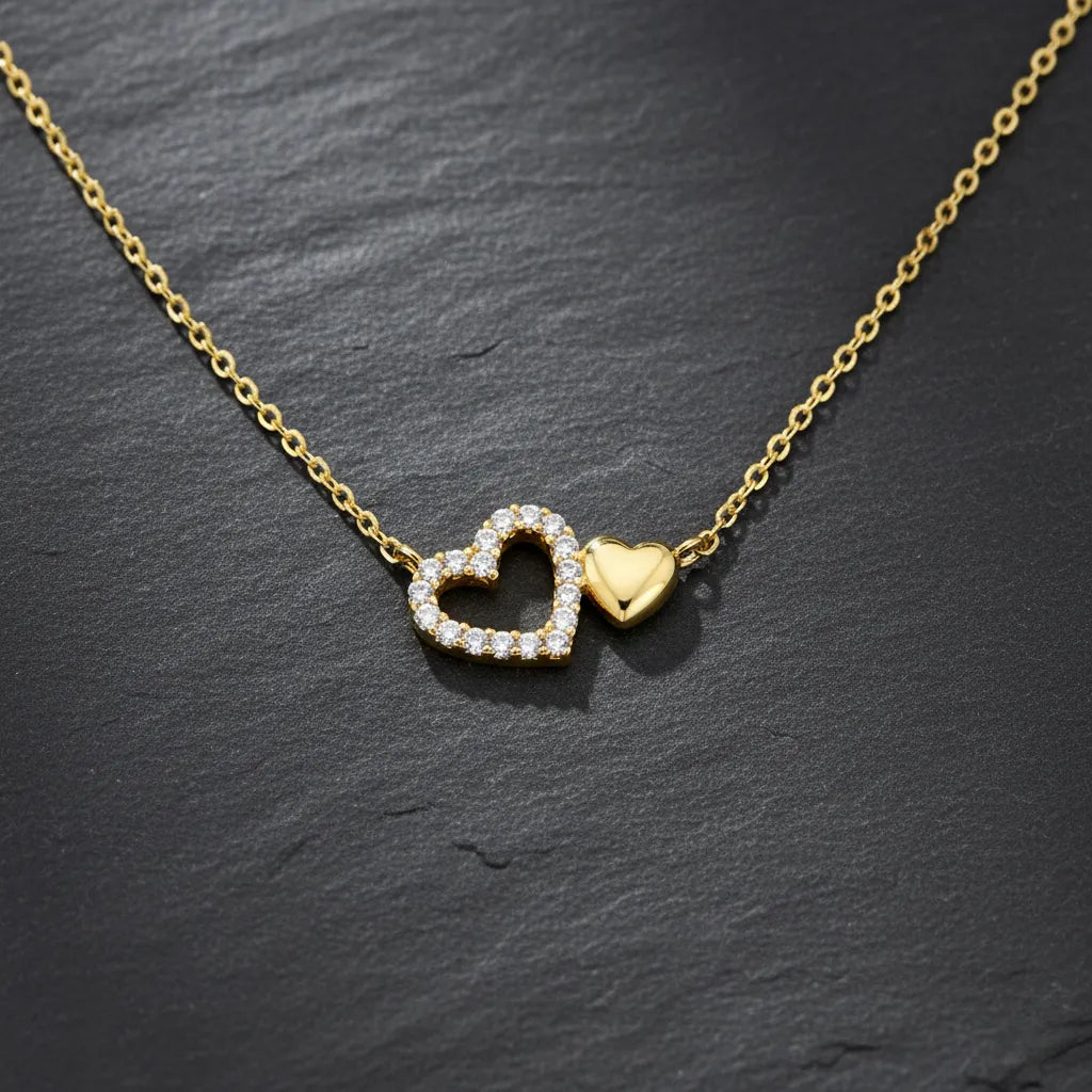 Vergoldete Damen Halskette mit zwei verbundenen Herzanhängern: ein pavé-besetztes Zirkonia-Herz und ein poliertes Goldherz, elegant präsentiert auf schwarzem Schieferhintergrund.
