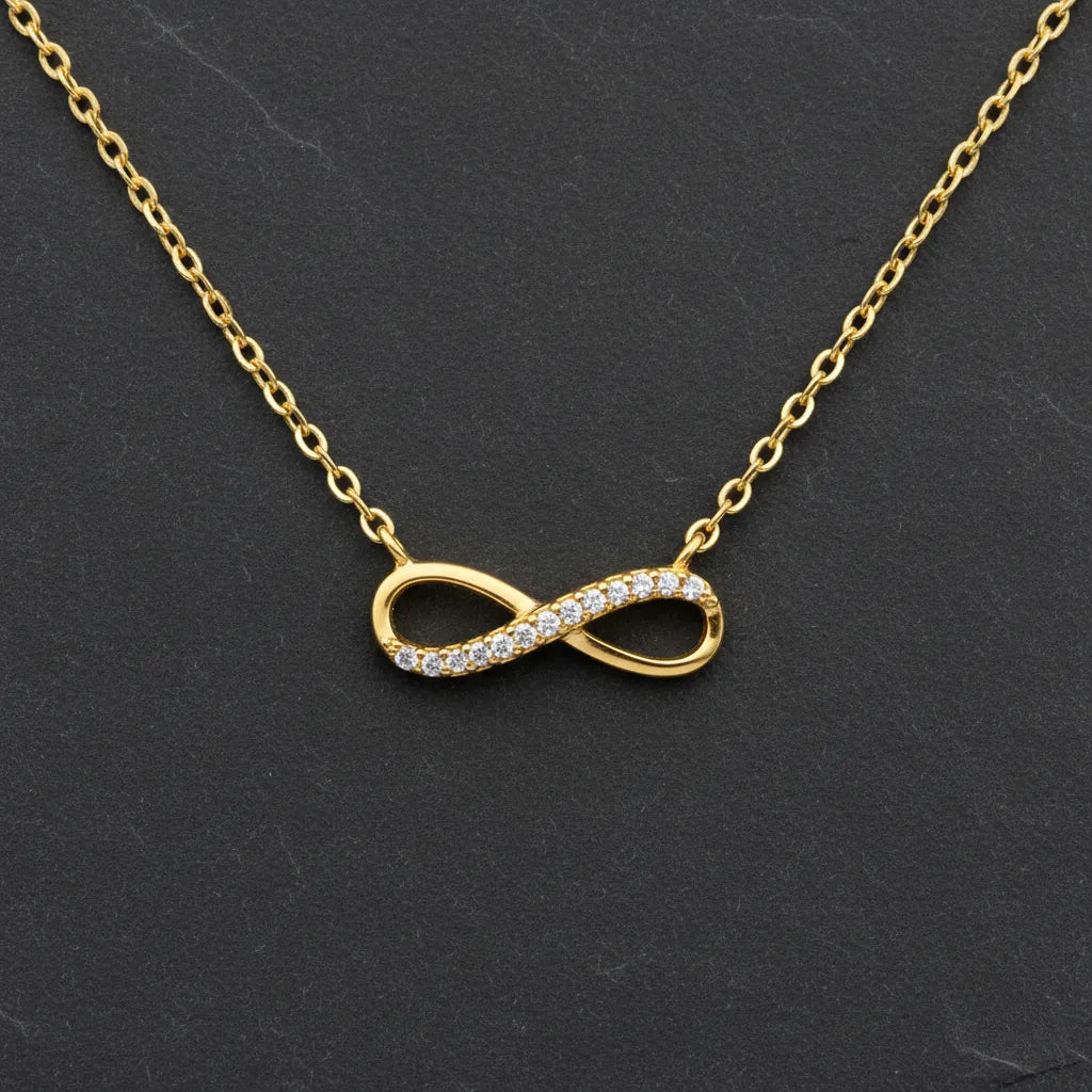 Vergoldete Damen Halskette mit filigranem Infinity-Anhänger, teilweise besetzt mit klar funkelnden Zirkonia-Steinen, elegant präsentiert auf dunklem Schieferhintergrund.
