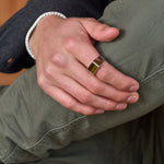 Herrenhand mit Tigerauge-Siegelring aus Edelstahl und geflochtenem Armband, lässiges Outfit (grüne Hose, dunkler Ärmel), natürliche Beleuchtung, Nahaufnahme.
