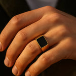 Herren-Siegelring aus vergoldetem Edelstahl mit rechteckigem schwarzem Stein, getragen am Ringfinger im Golden-Hour-Licht, Nahaufnahme.
