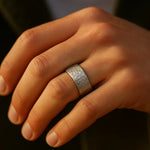 Herrenring aus Edelstahl, gehämmerte Oberfläche, breite Schiene – getragen am Ringfinger; Nahaufnahme im warmen Fensterlicht, natürliche Hauttöne.
