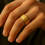 Herrenring vergoldeter Edelstahl, 8 mm breit, gehämmerte Oberfläche – getragen am Ringfinger; Nahaufnahme im warmen Fensterlicht, natürliche Hauttöne.
